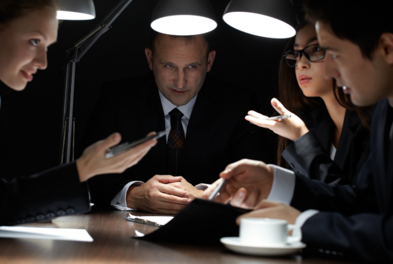 Image is of a group of people talking at a table with a look of secrecy, concept of things insurance companies don't want you to know