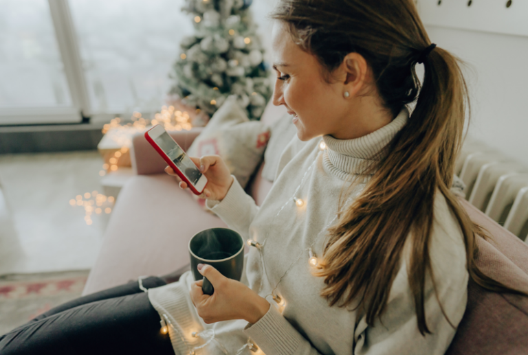 Image is of a woman sitting in her living room while shopping online from her phone with a Christmas tree in the background, concept of cyber crime and identity theft during the holiday shopping season