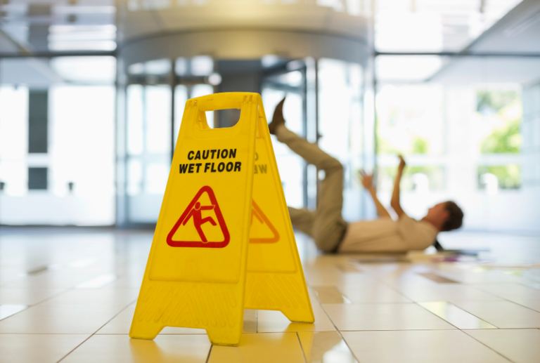 Image is of closeup of a yellow floor sign that reads 'caution wet floor' and in the background shows a man slipping and falling, concept of what to do if injured at a store or business