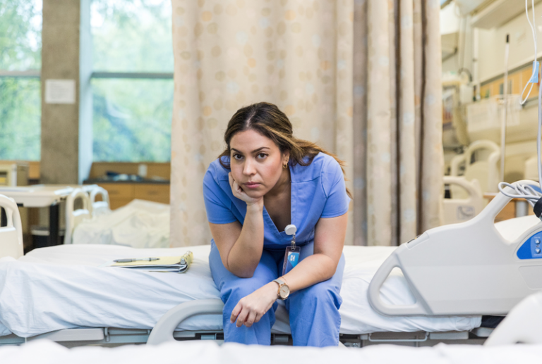 Image is of a nurse sitting on the side of an empty hospital bed resting her head on her hand and looking upset, concept of what to do if a complaint is filed against your nursing license.