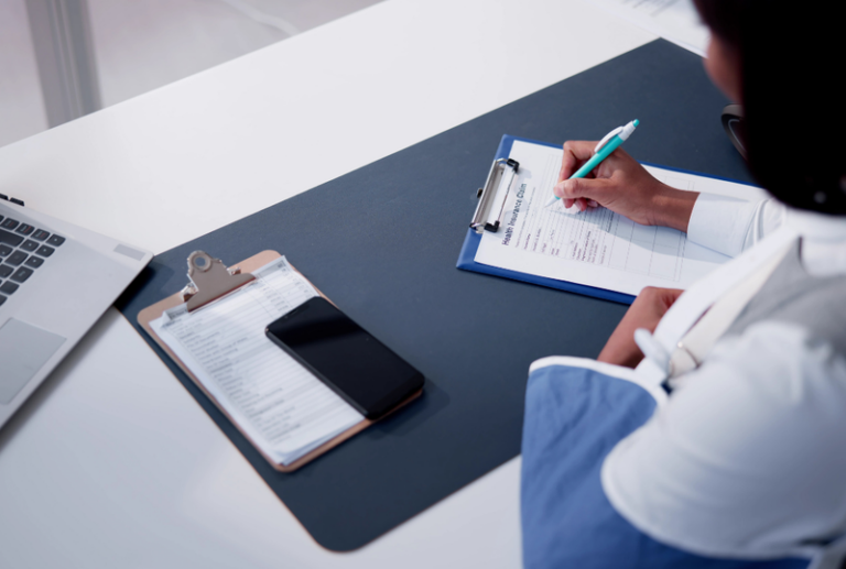 Image is of a view from above a woman with one arm in a sling, and filling out papers at a desk, concept of what is covered by workers' compensation insurance