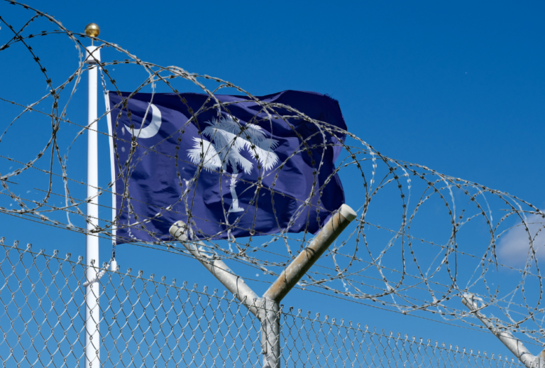 Image is of the South Carolina state flag behind a fence and barbed wire, concept of South Carolina's return to firing squad and capital punishment