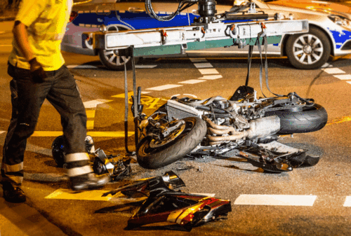 Image is of a wrecked motorcycle on the road with a road worker cleaning the road of the debris, concept of what to do after a hit and run motorcycle accident in Aiken.
