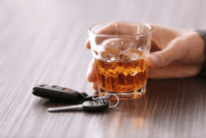 Image is of a person's hand holding a glass of whiskey and a set of keys is laying next to the glass, concept of a first-time DUI in Aiken