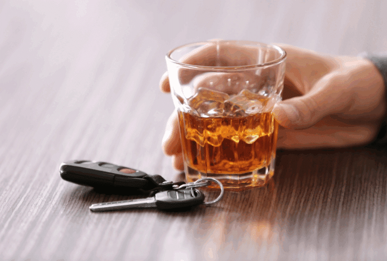 Image is of a person's hand holding a glass of whiskey and a set of keys is laying next to the glass, concept of a first-time DUI in Aiken