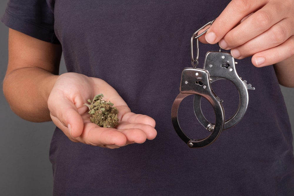 Image shows a person holding marijuana buds in one hand and handcuffs in the other, representing how state drug charges begin with simple possession investigations.