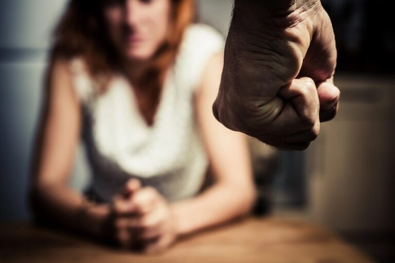 Image shows a raised clenched fist and a distressed individual seated nearby, representing domestic violence offenses and the serious criminal accountability under South Carolina law.