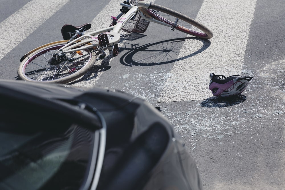Image shows a damaged bicycle lying on a crosswalk with a helmet and shattered glass nearby, concept of gathering evidence to prove fault in a bike accident.