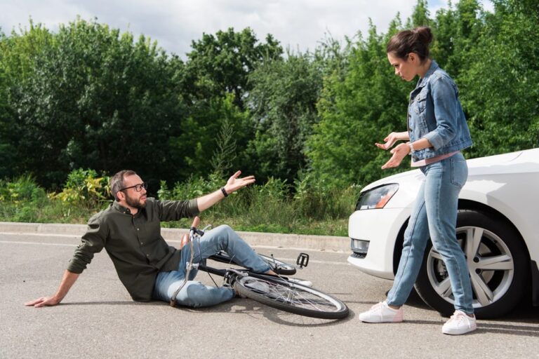 Image shows a man sitting on the road beside his bicycle after a collision with a car while the driver speaks to him, illustrating how fault in a bike accident may be determined.