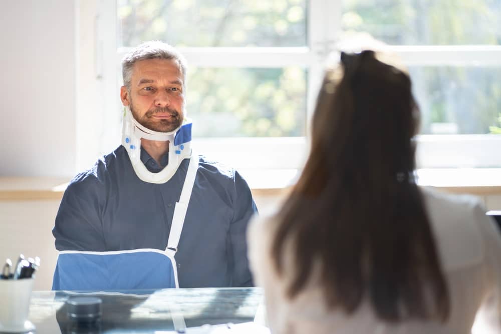 Image is of an injured motorcyclist wearing a neck brace and arm sling while speaking with a medical professional, showing recovery from injuries caused by motorcycle accidents.