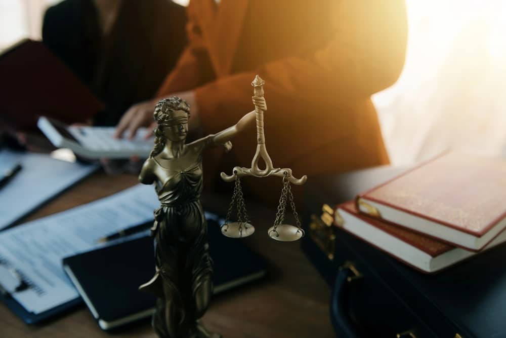 Image shows a Lady Justice statue on a desk while lawyers review documents in the background, symbolizing legal issues that arise after truck accidents in SC.