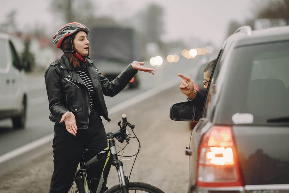 Image shows a cyclist gesturing toward a car driver after a roadway conflict, highlighting fault disputes and safety risks in bike accidents