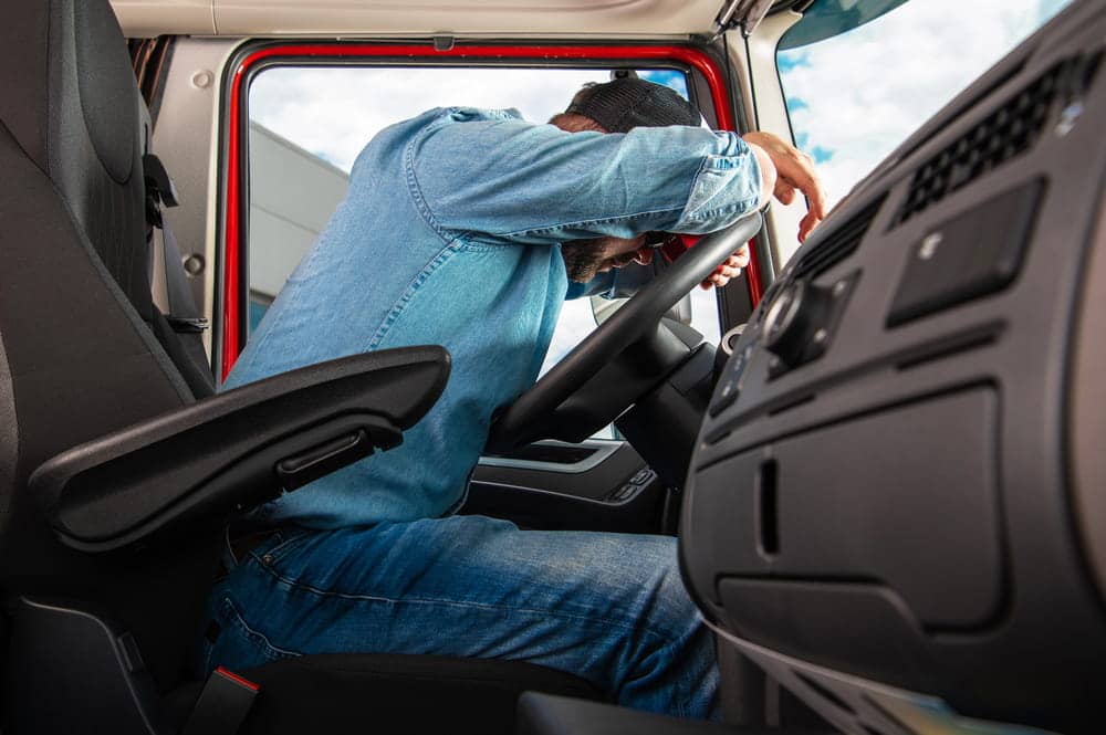 Image is of a tired truck driver resting on the steering wheel inside the cab, illustrating driver fatigue and how to sue a trucking company after a serious crash.