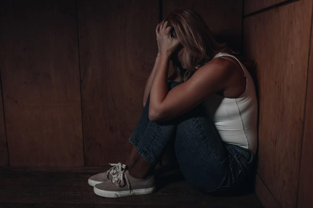 Image is of a woman sitting on the floor holding her head, representing emotional impact linked to domestic violence convictions