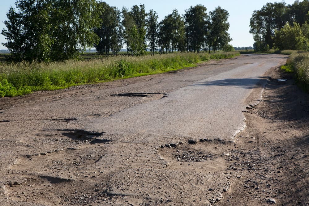 Image is of a damaged rural roadway with multiple potholes and uneven surfaces, highlighting poor road conditions in Aiken that may lead to accidents