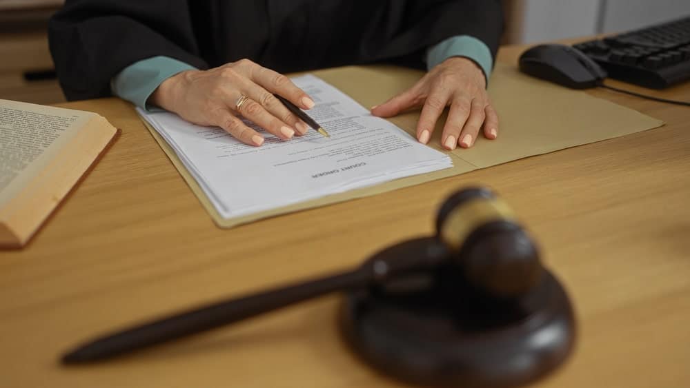 Image is of a person signing legal paperwork with a judge’s gavel nearby, representing compliance requirements under a protective order