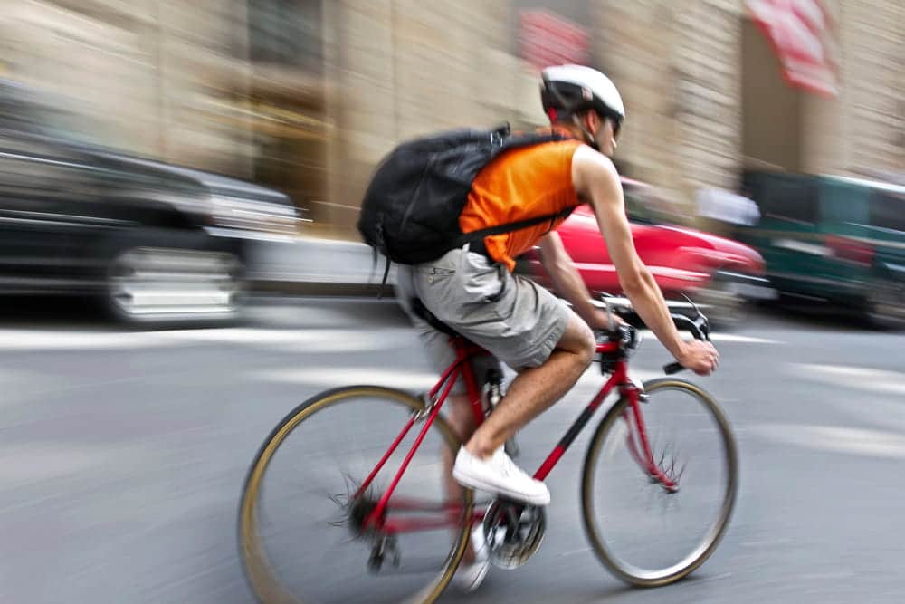 Image is of a cyclist riding through urban traffic with cars nearby, concept of roadway risks and safety while biking in Aiken.