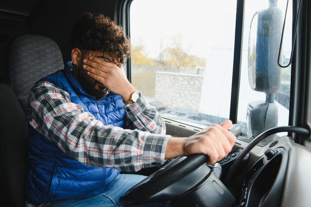 Image is of a tired truck driver holding his face while driving, representing driver fatigue linked to truck accidents in Aiken