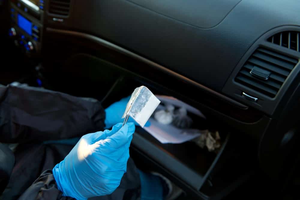 Image is of a police officer using gloves to collect a white substance from a car glove compartment, illustrating drug possession in Aiken investigation