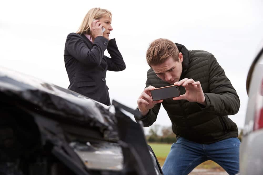 Image is of a man photographing vehicle damage after a collision while another person makes a phone call, concept of a car accident lawsuit involving crash scene documentation and evidence collection.