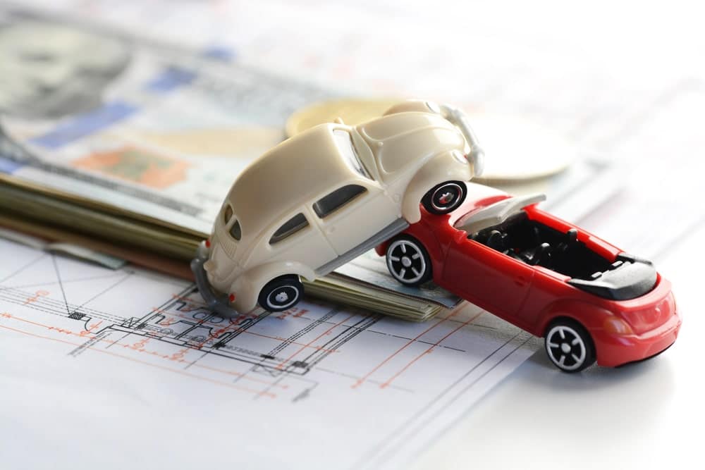 Image is of two miniature cars involved in a collision placed over financial paperwork, concept of a car accident lawsuit involving insurance claims and compensation disputes.