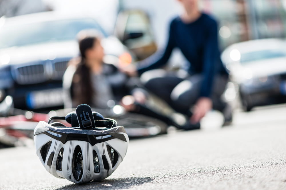 Image is of a bicycle helmet lying on the road near a damaged bicycle after a collision, concept of a bicycle accident lawyer helping injured cyclists pursue financial recovery.