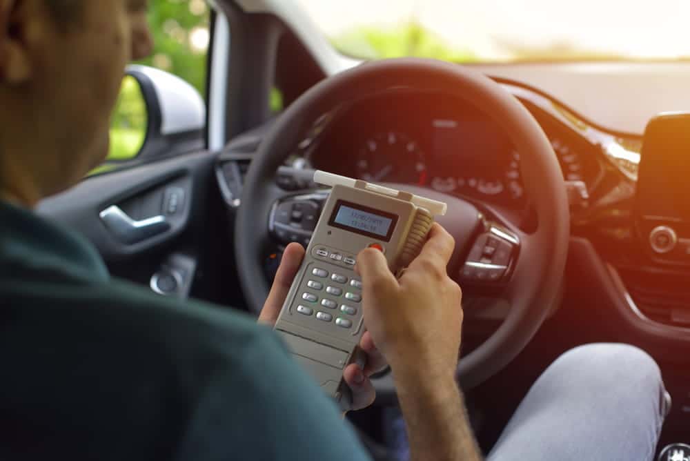 Image is of a driver holding a breath testing device inside a vehicle, concept of a DUI license suspension lawyer addressing chemical testing issues.