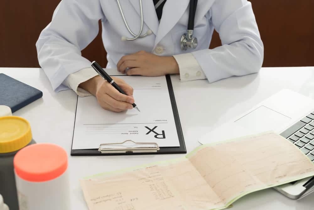 Image is of a doctor writing a prescription while reviewing patient records, concept of an Aiken defective drugs lawyer evaluating a medication injury claim.