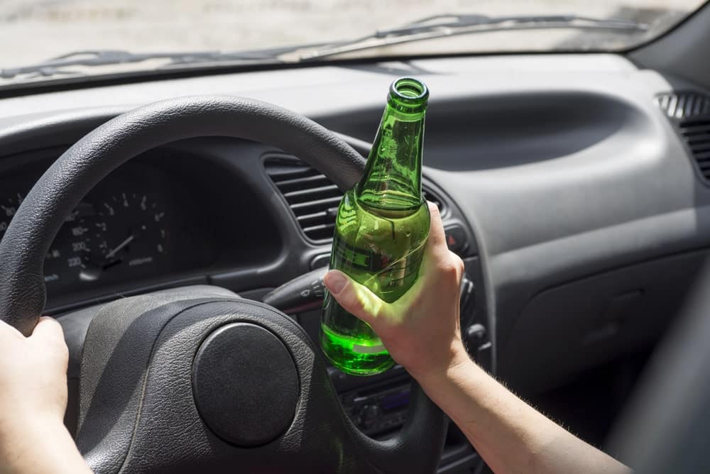 Image is of a driver holding an alcoholic beverage while operating a vehicle, concept of a first offense DUI lawyer representing individuals accused of impaired driving offenses.