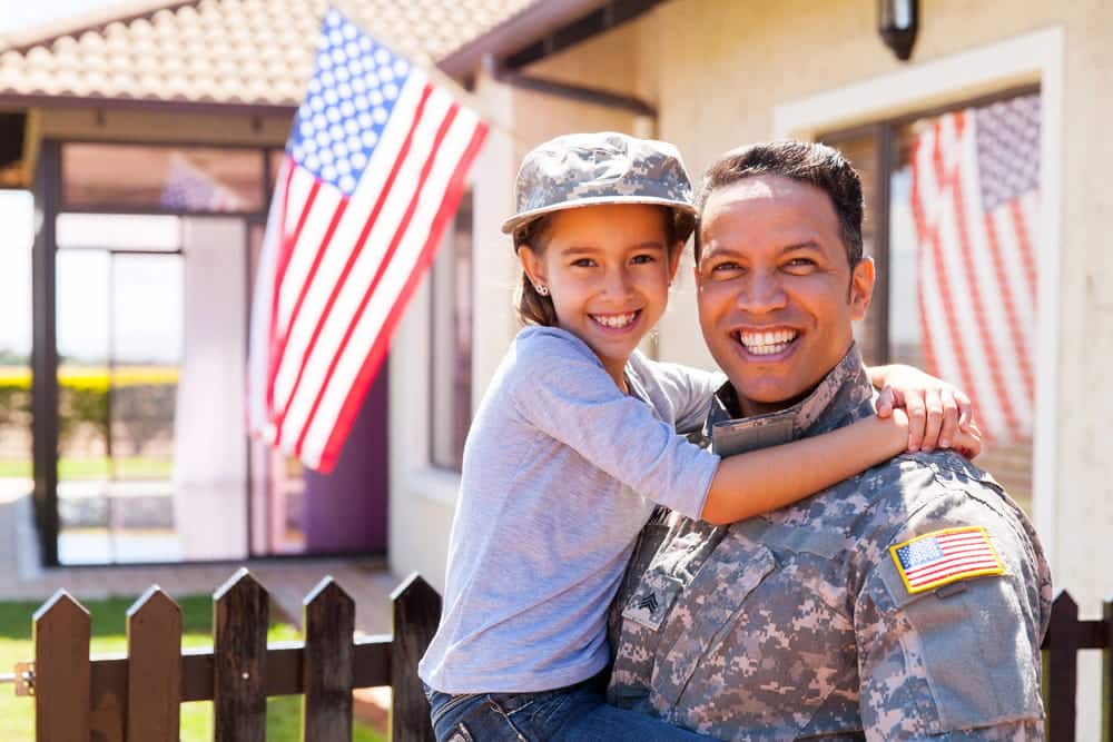 Image is of a military service member holding a child outside a family home, concept of custody and parenting concerns handled by a military divorce lawyer in Aiken.