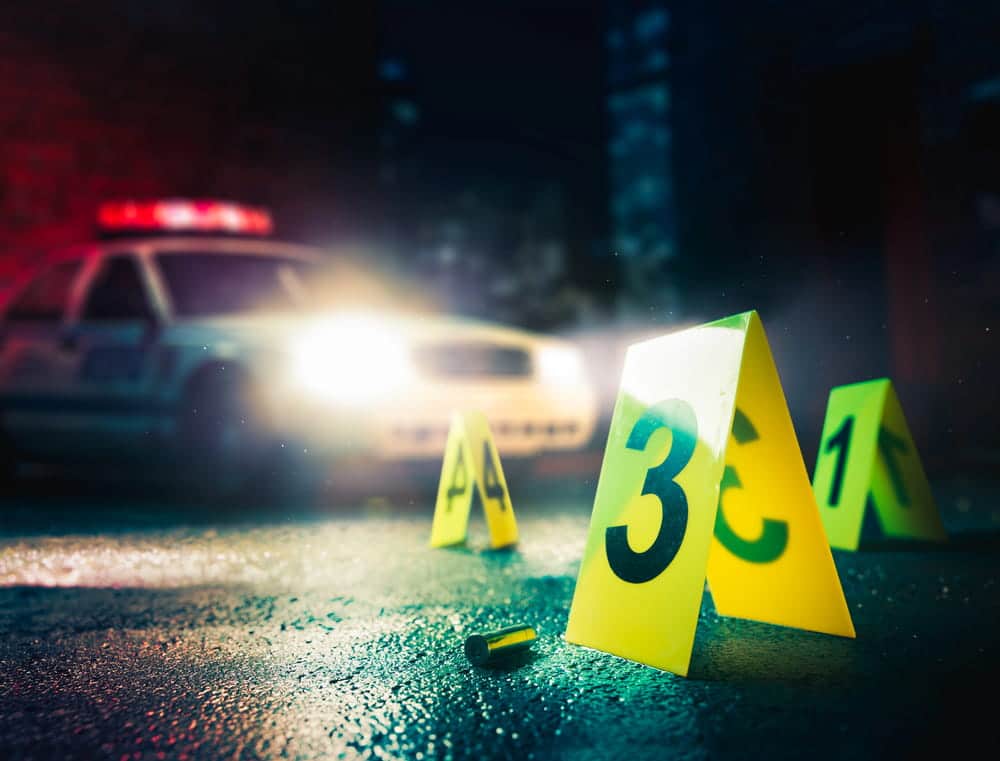 Image is of evidence markers and a bullet casing at a crime scene with a police vehicle in the background, concept of an Aiken murder defense lawyer reviewing forensic evidence in a criminal case.