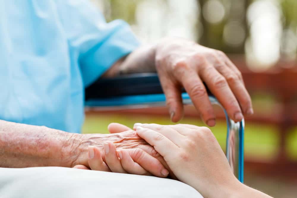 Image is of a caregiver holding an elderly person's hand while they sit in a wheelchair, concept of an Aiken nursing home abuse and neglect lawyer advocating for victims of neglect and abuse.