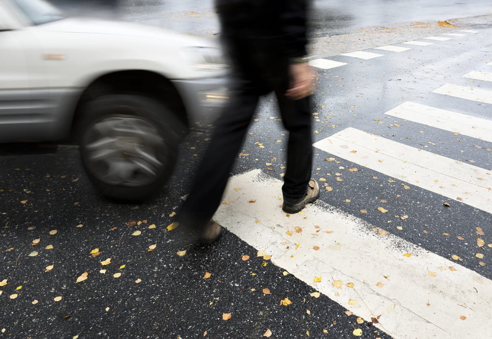 Image is of a pedestrian crossing a roadway near moving traffic, concept of a pedestrian accident lawyer evaluating crosswalk collision cases.