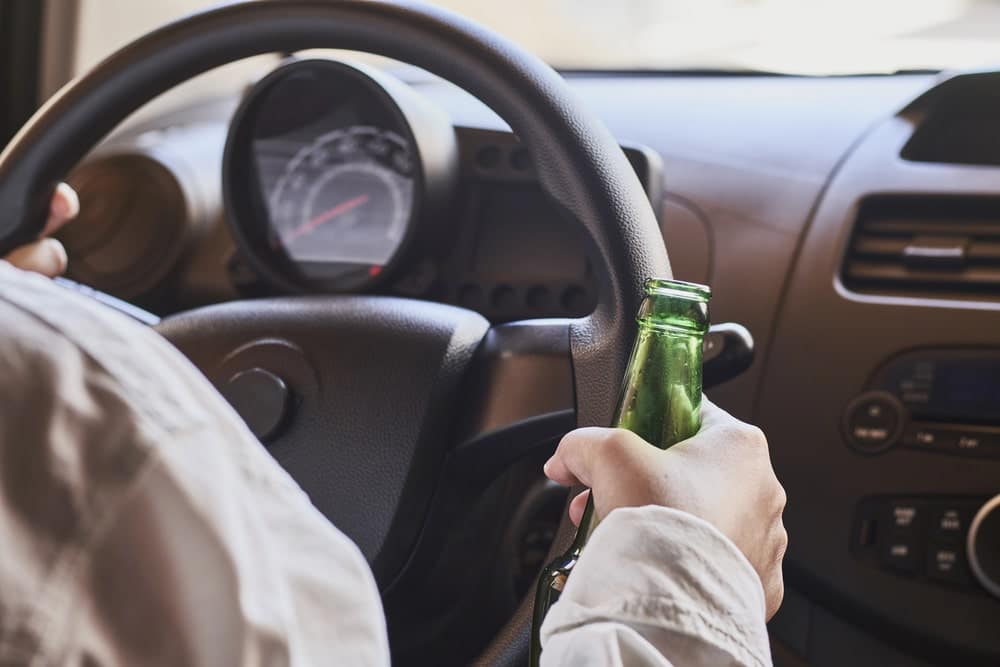 Image is of a driver holding an alcoholic beverage while operating a vehicle, concept of a DUI lawyer in Aiken handling drunk driving allegations.