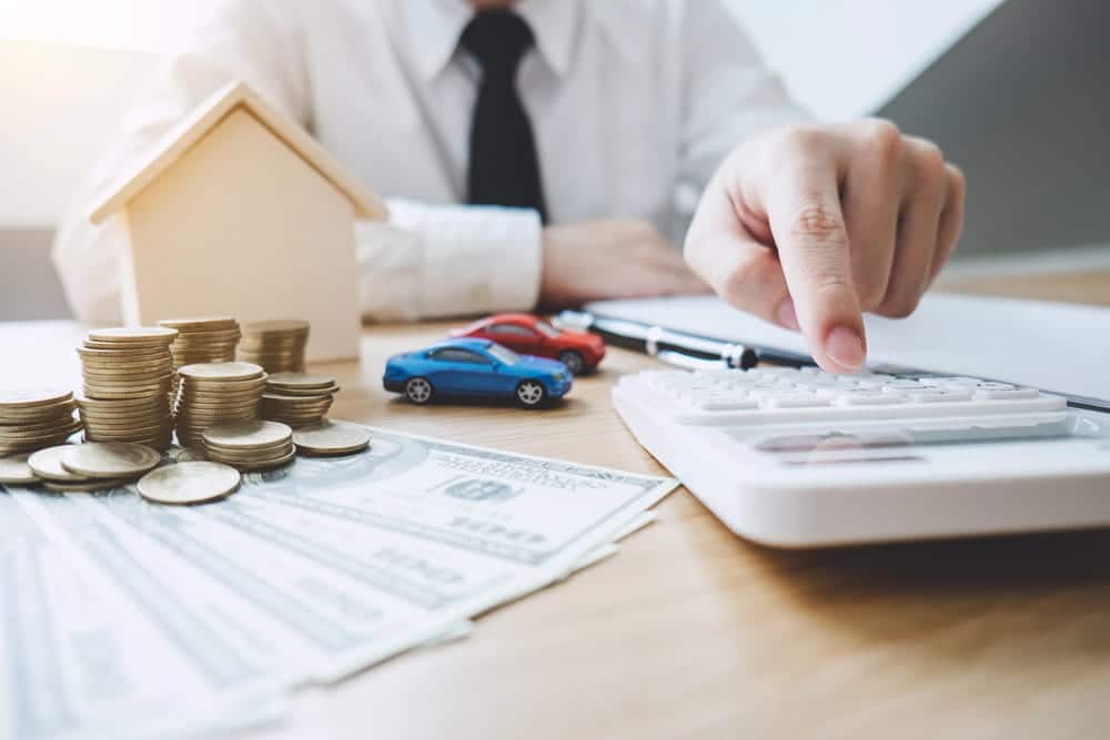 Image is of a person calculating finances beside money, cars, and a model home, concept of how property divided during divorce.