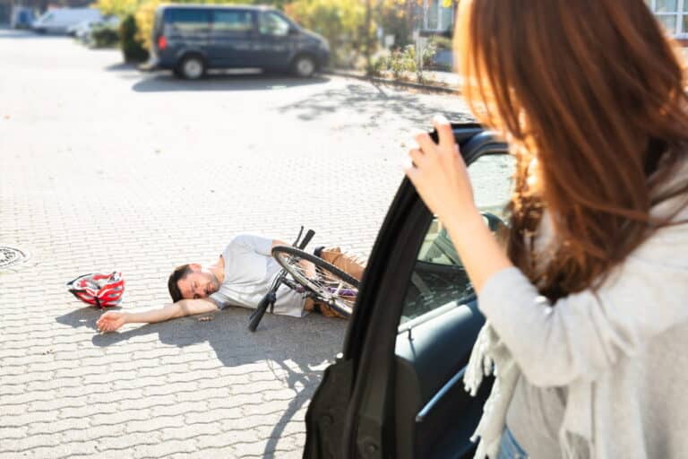 Image is of a cyclist injured beside a car while a driver looks on, concept of an accident while biking in Aiken.
