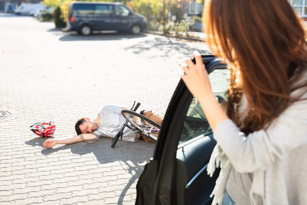 Image is of a cyclist injured beside a car while a driver looks on, concept of an accident while biking in Aiken.
