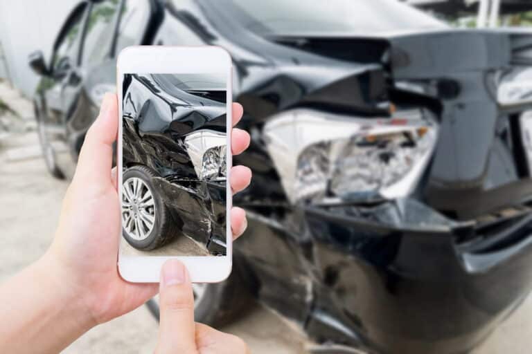 Image is of a person photographing damage to a black car using a smartphone, concept of documenting vehicle damage after a hit-and-run in Aiken.