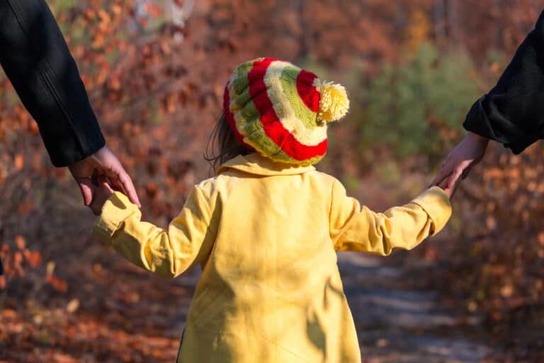 Image is of a child walking outdoors while holding both parents' hands, illustrating shared parenting time under joint custody arrangements.