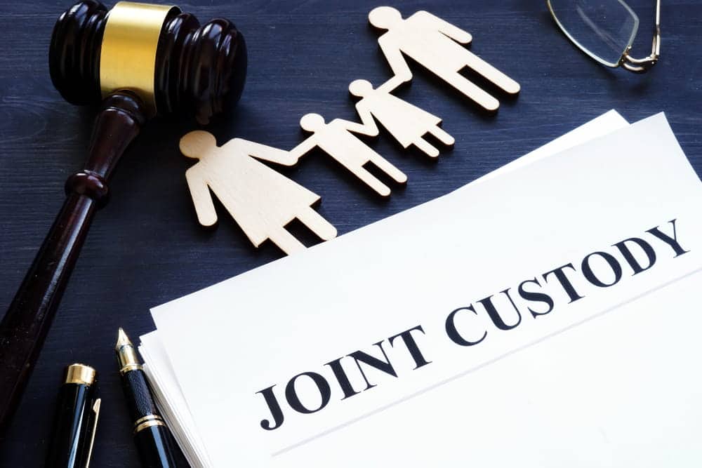 Image is of joint custody documents alongside a judge's gavel and wooden family figures, representing legal arrangements for shared parental responsibilities.