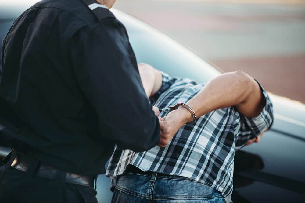 Image is of a police officer handcuffing a suspect beside a vehicle, illustrating how traffic stops can lead to drug arrests in Aiken.