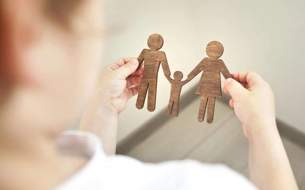 Image is of hands holding wooden figures representing two parents and a child, concept of eligibility to adopt a child in South Carolina.